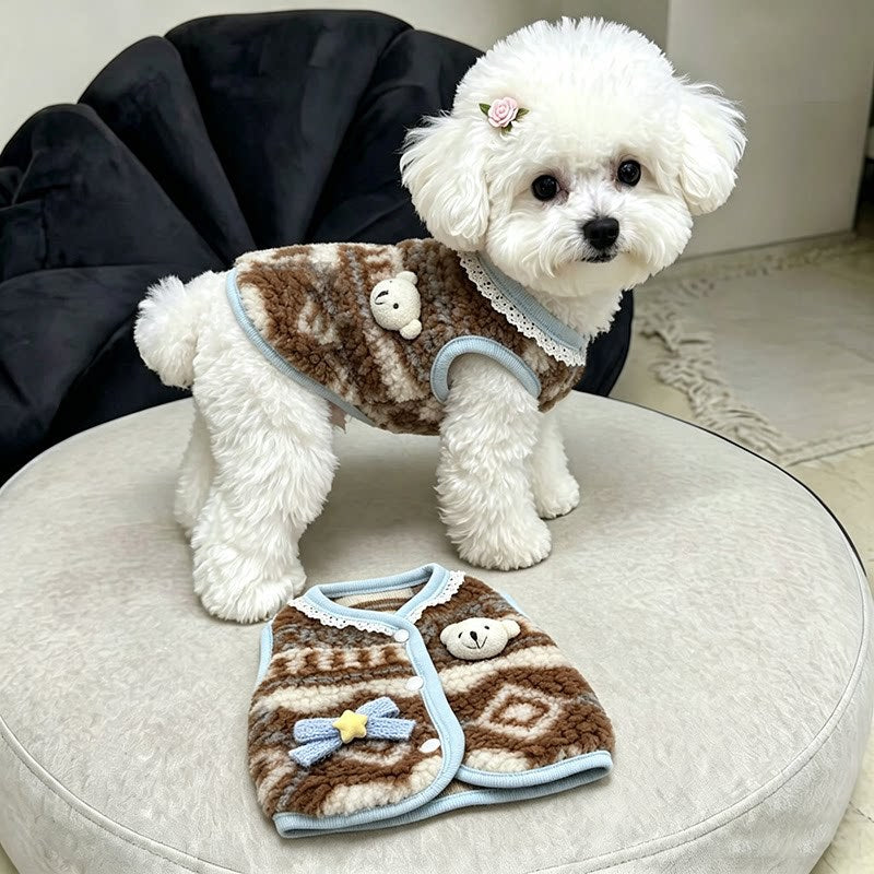 A small white fluffy dog stands on a round beige cushion wearing a brown and beige patterned pet sweater with light blue trim, while a matching folded sweater lies in front. A black chair is visible in the background.