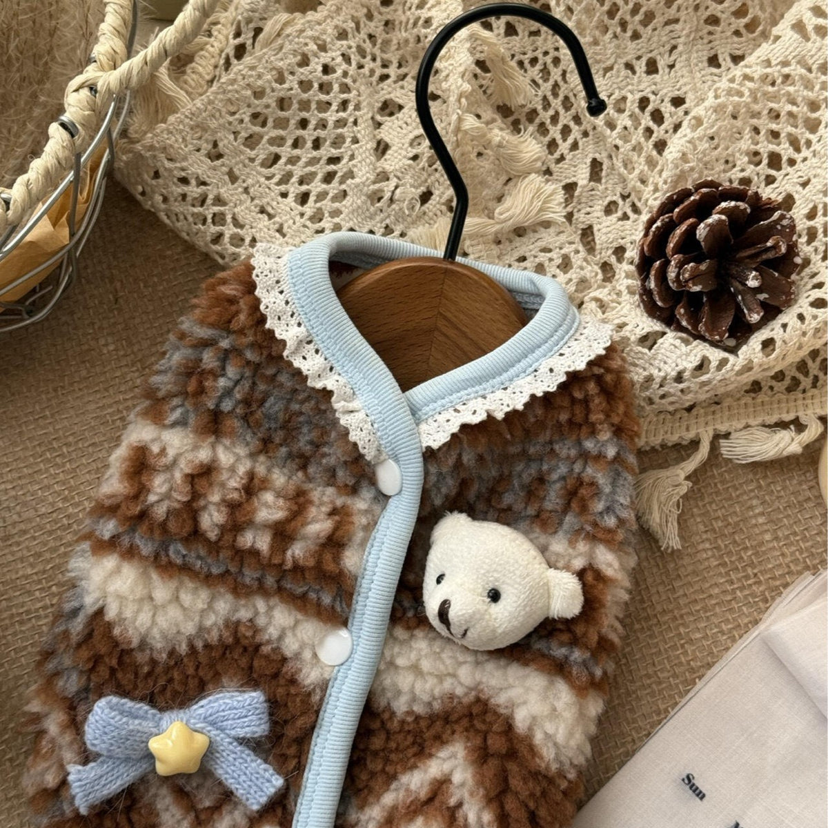 A pet cardigan vest with brown, beige, and gray striped fuzzy fabric, light blue trim, a lace collar, a small plush bear face, and a blue bow with a yellow star, displayed on a wooden hanger.