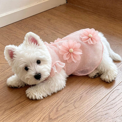A small white dog lying on a wooden floor wearing a pink mesh pet dress with large flower appliqués.