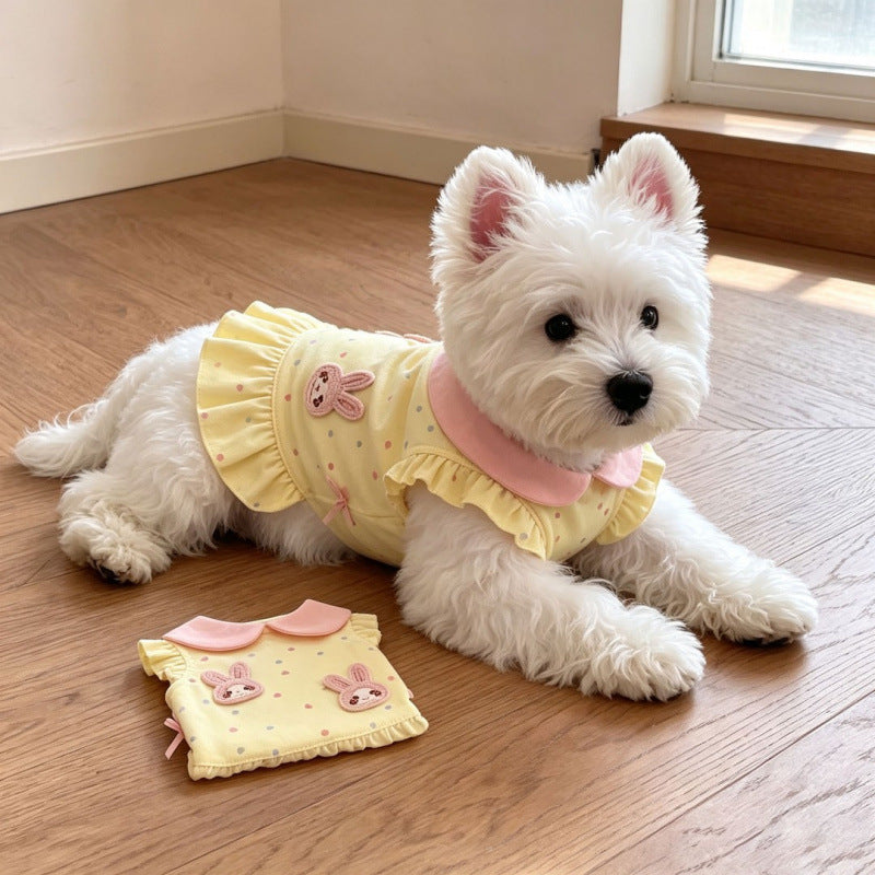 A small white dog lies on a wooden floor wearing a pale yellow pet top with pink ruffles and bunny graphics. A matching pet top is placed on the floor beside it.