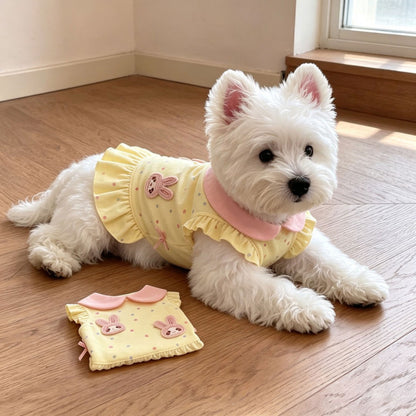 A small white dog lies on a wooden floor wearing a pale yellow pet top with pink ruffles and bunny graphics. A matching pet top is placed on the floor beside it.