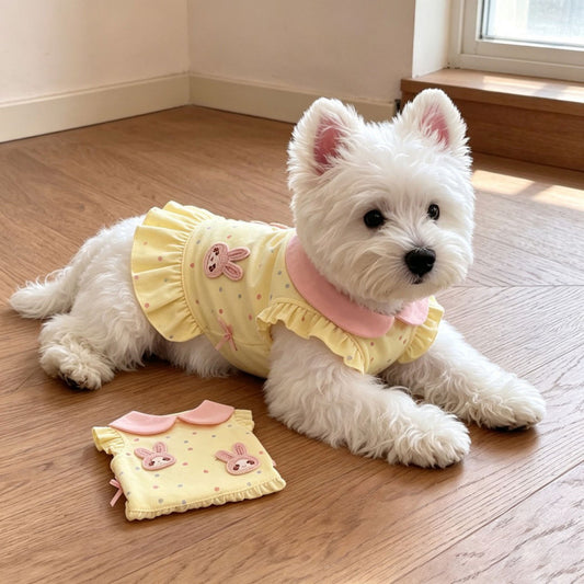 A small white dog lies on a wooden floor wearing a pale yellow pet top with pink ruffles and bunny graphics. A matching pet top is placed on the floor beside it.