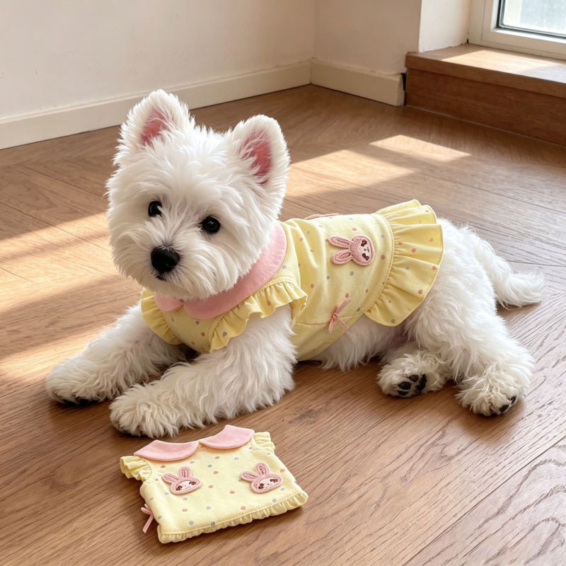 A small white dog lies on a wooden floor wearing a pale yellow ruffled pet dress with pink trim and bunny graphics. A matching folded version of the dress sits in front of the dog.