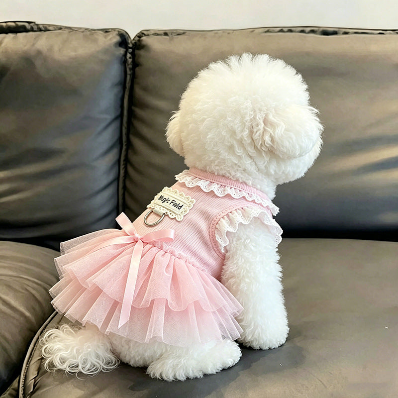 A small white dog sits on a gray leather couch wearing a pink ribbed pet dress with a ruffled collar, lace trim, and a layered tulle skirt. The back of the dress has a decorative patch that reads 'best friend'.