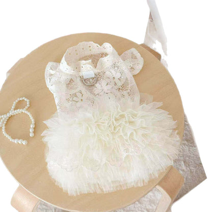 A white lace and ruffle pet dress displayed on a round wooden stand, accompanied by a matching white pearl headband.