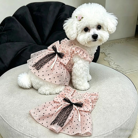 A small white fluffy dog sits on a round light gray cushion wearing a pink polka-dot pet dress with black lace trim and a bow. The matching dress is laid flat in front on the cushion.