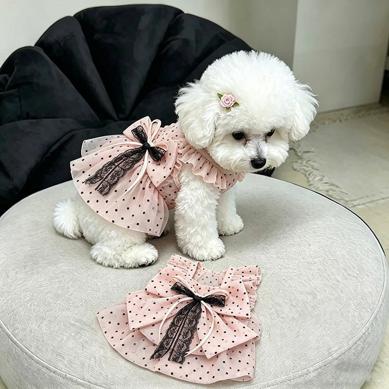 A small white fluffy dog wearing a pink polka-dot pet dress with a large ruffled bow and lace trim, sitting on a round light gray cushion with the dress laid flat in front.