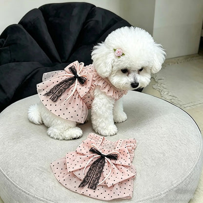 A small white fluffy dog wearing a pink polka-dot pet dress with a large ruffled bow and lace trim, sitting on a round light gray cushion with the dress laid flat in front.